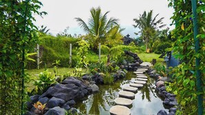 Un sendero de jardín con piedras para pisar que lleva a una fuente de agua rodeada de abundante vegetación y palmeras.
