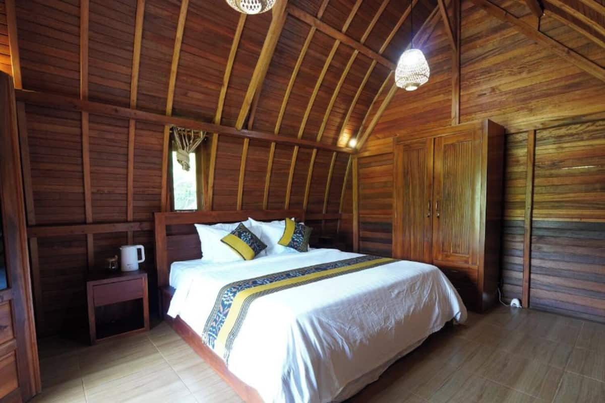 Una habitación con piso de madera, una cama, una mesita de noche, un armario y una lámpara colgante.