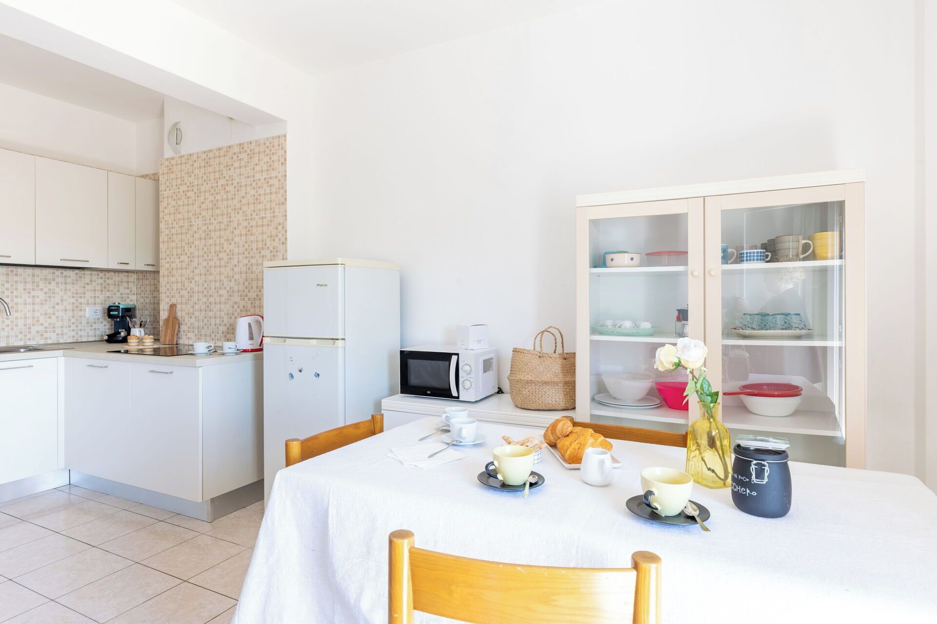 Una cocina compacta con refrigerador, microondas y cafetera. Hay una mesa de comedor con mantel blanco, una cesta y un tarro.