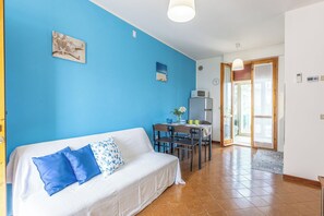 Ein Zimmer mit blauer Wand, einem weißen Sofa mit blau-weißen Kissen, einem Esstisch mit Stühlen, einer Mikrowelle und einer Uhr an der Wand.