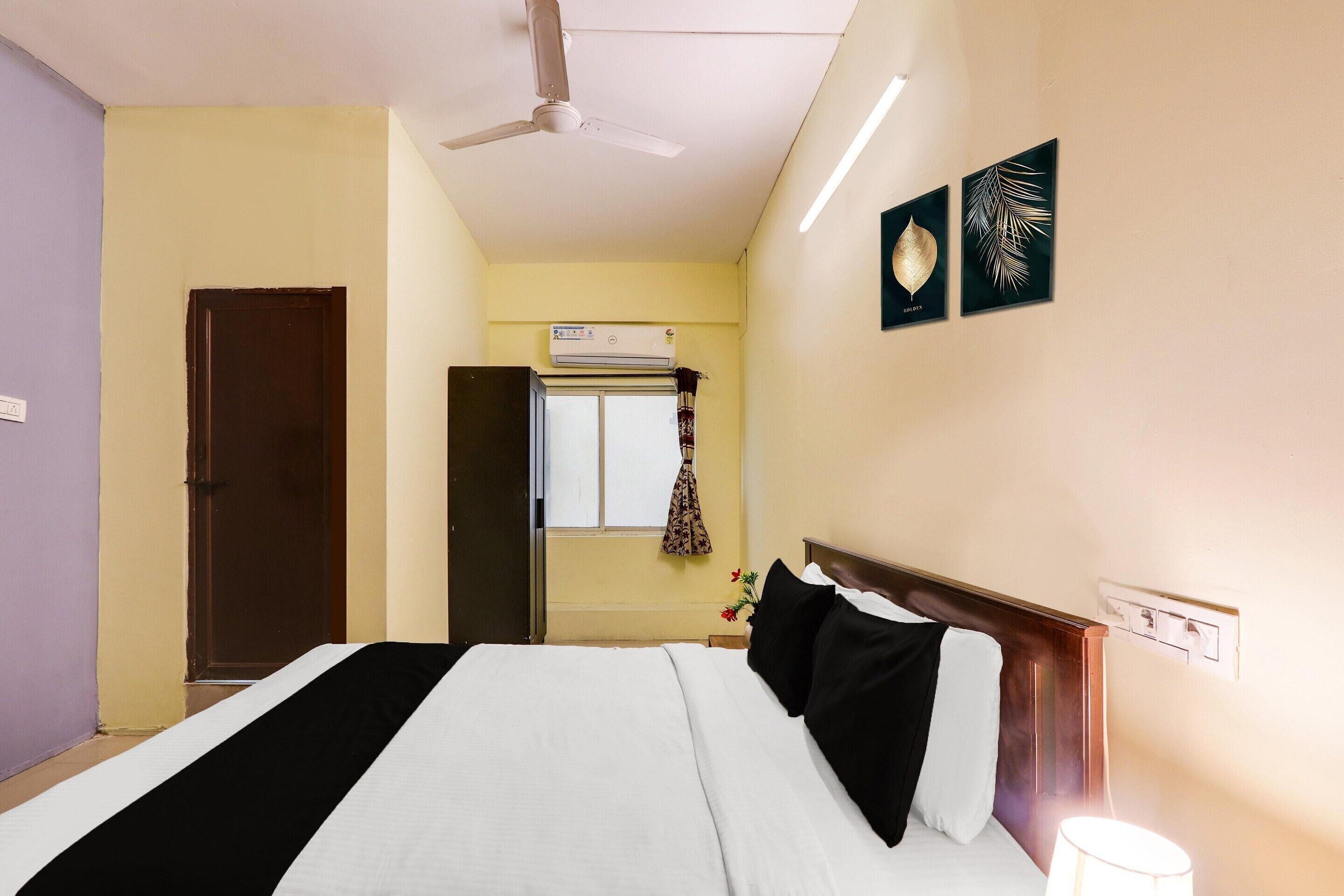 A hotel room with a bed, a ceiling fan, an air conditioning unit, a window with a curtain, and wall-mounted artwork.
