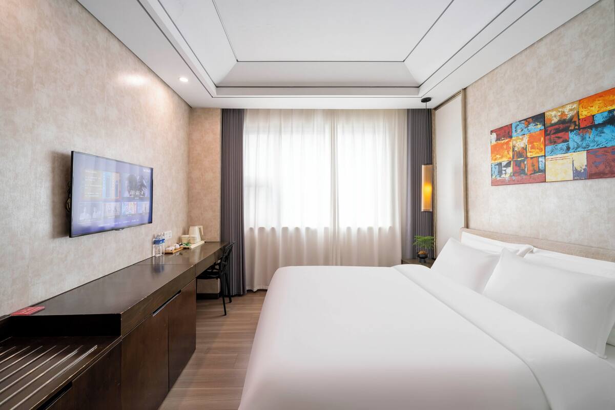 Habitación de hotel con cama, escritorio con televisor, ventana con cortinas y un cuadro en la pared.