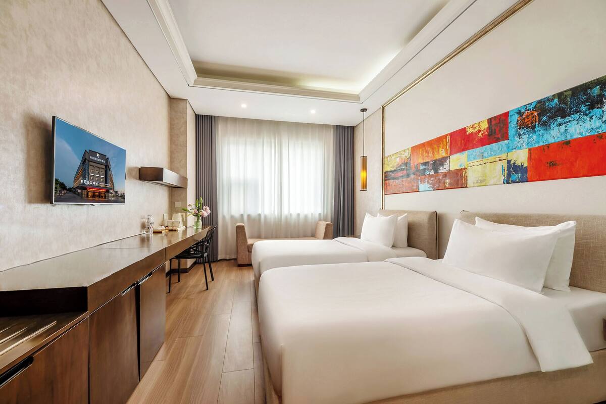Habitación de hotel con dos camas, un escritorio, una silla, un televisor y un cuadro abstracto grande.