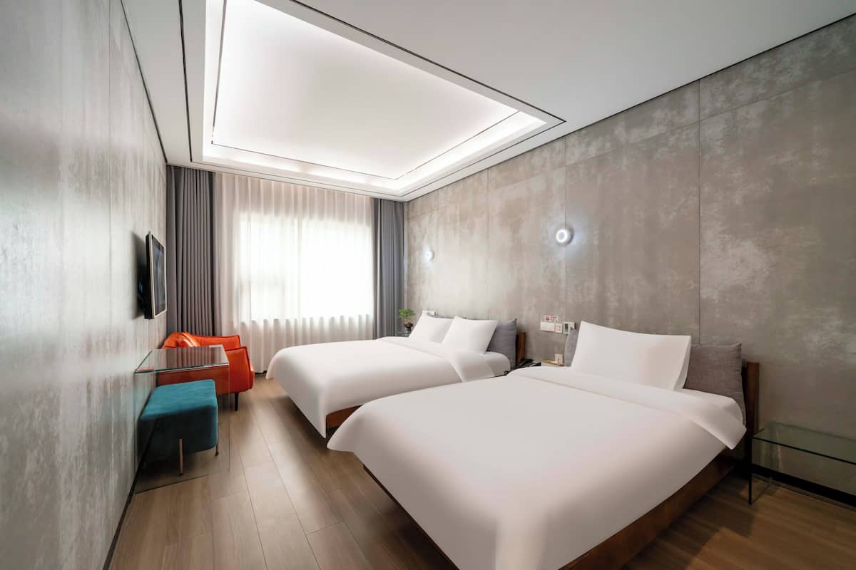 Una habitación de hotel moderna con dos camas, una mesa de vidrio y una zona de estar con cojines naranjas.