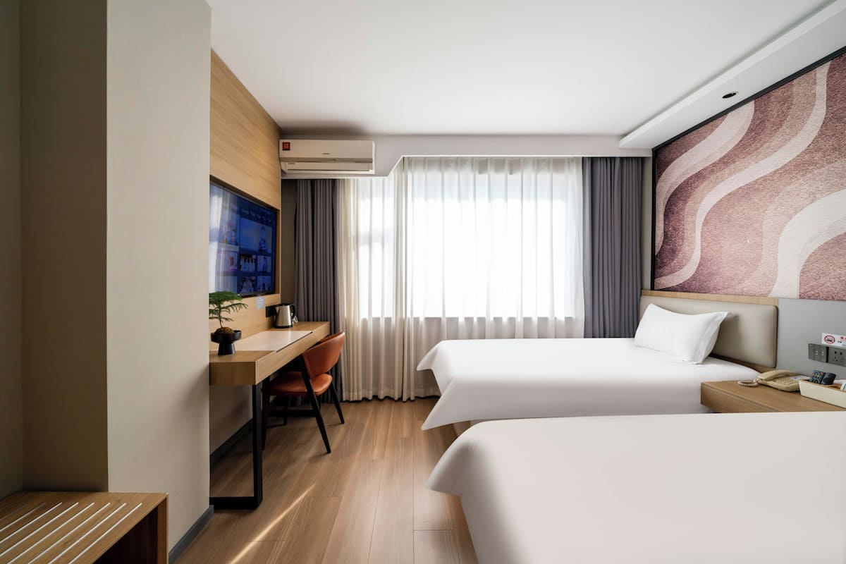 Habitación de hotel con dos camas, un escritorio, una silla, un televisor y un ventanal grande con cortinas.