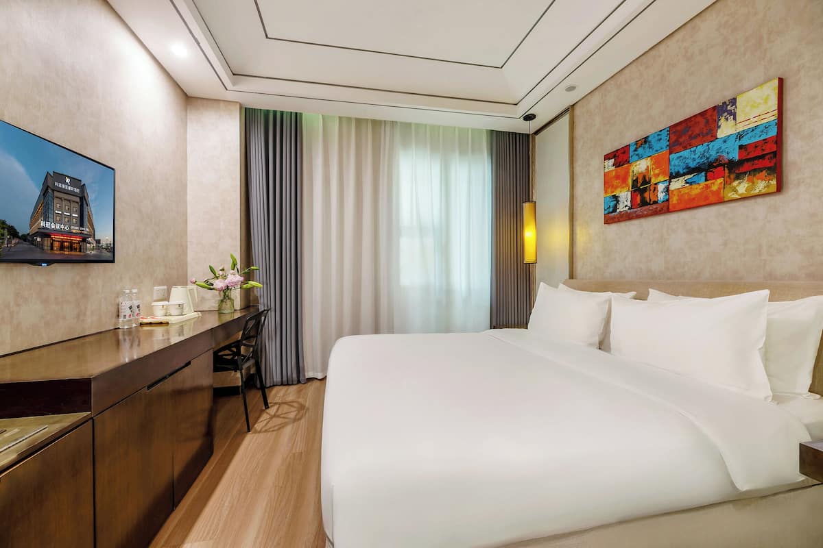 Habitación de hotel con cama, escritorio, silla, televisor, un cuadro y ventana con cortinas.