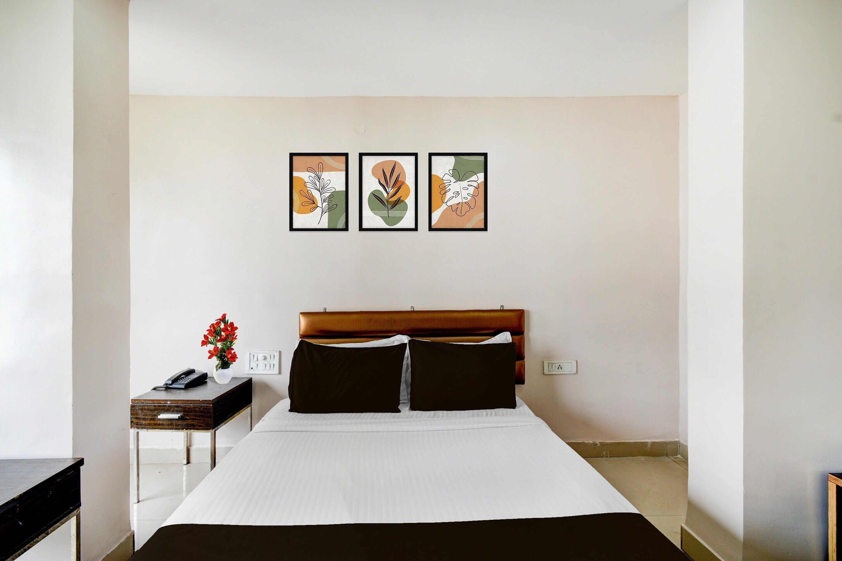 A hotel room with a bed, a bedside table with a phone and vase, and three framed wall art pieces.