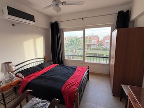 A bedroom with a bed, a desk, a wardrobe, a ceiling fan, and a window with a view of buildings.