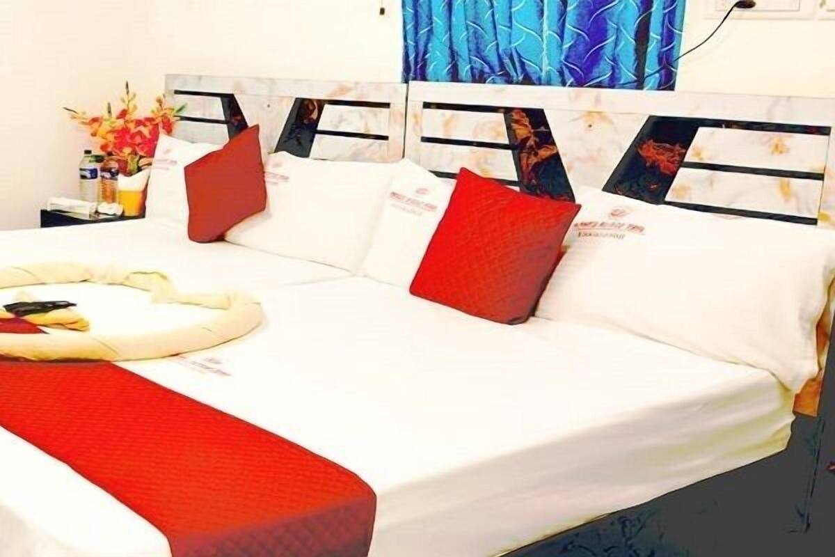 A hotel room with a bed, pillows, a red blanket, a nightstand with a vase of flowers, and a headboard with a patterned design.