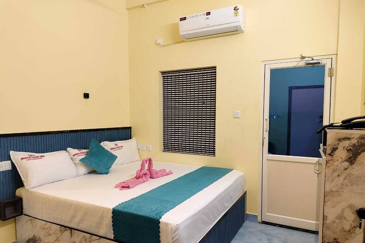 A hotel room with a bed, a window with blinds, an air conditioning unit, and a door.