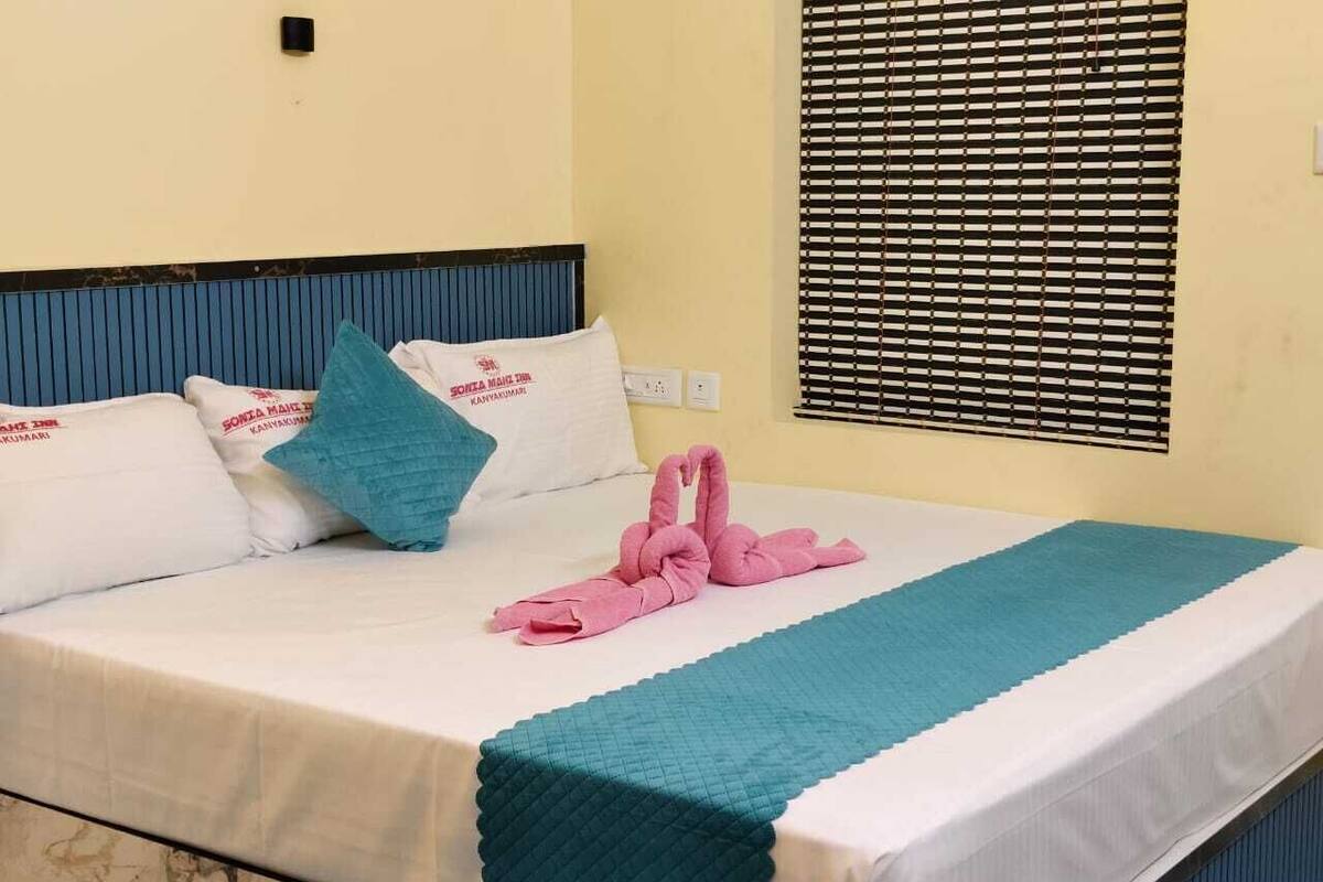 A hotel room with a bed, pillows, a window with blinds, and a wall-mounted light switch.