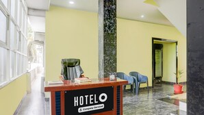 A reception desk with a 'HOTELO' sign, a vertical pillar, and a hallway with chairs and a potted plant.