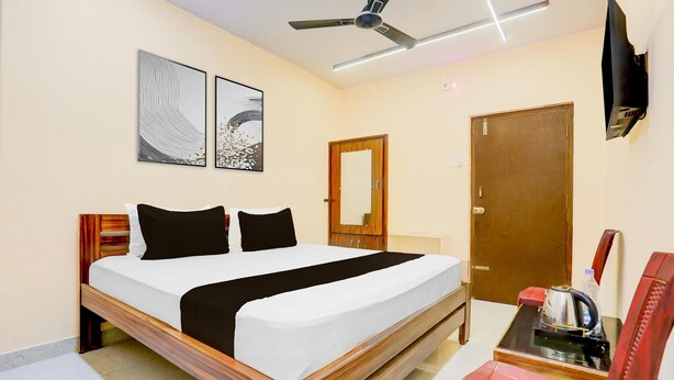 A hotel room with a bed, a desk with a kettle, a chair, a ceiling fan, and two wall-mounted artworks.