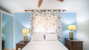 A bedroom with a bed, two nightstands with lamps, a ceiling fan, and a window with a view. - Garden Condos 9, Affordable APT in Sosua Downtown! (Sosúa)