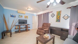 A living room with a television, a brown armchair, a coffee table, and a sofa. - Garden Condos 9, Affordable APT in Sosua Downtown! (Sosúa)