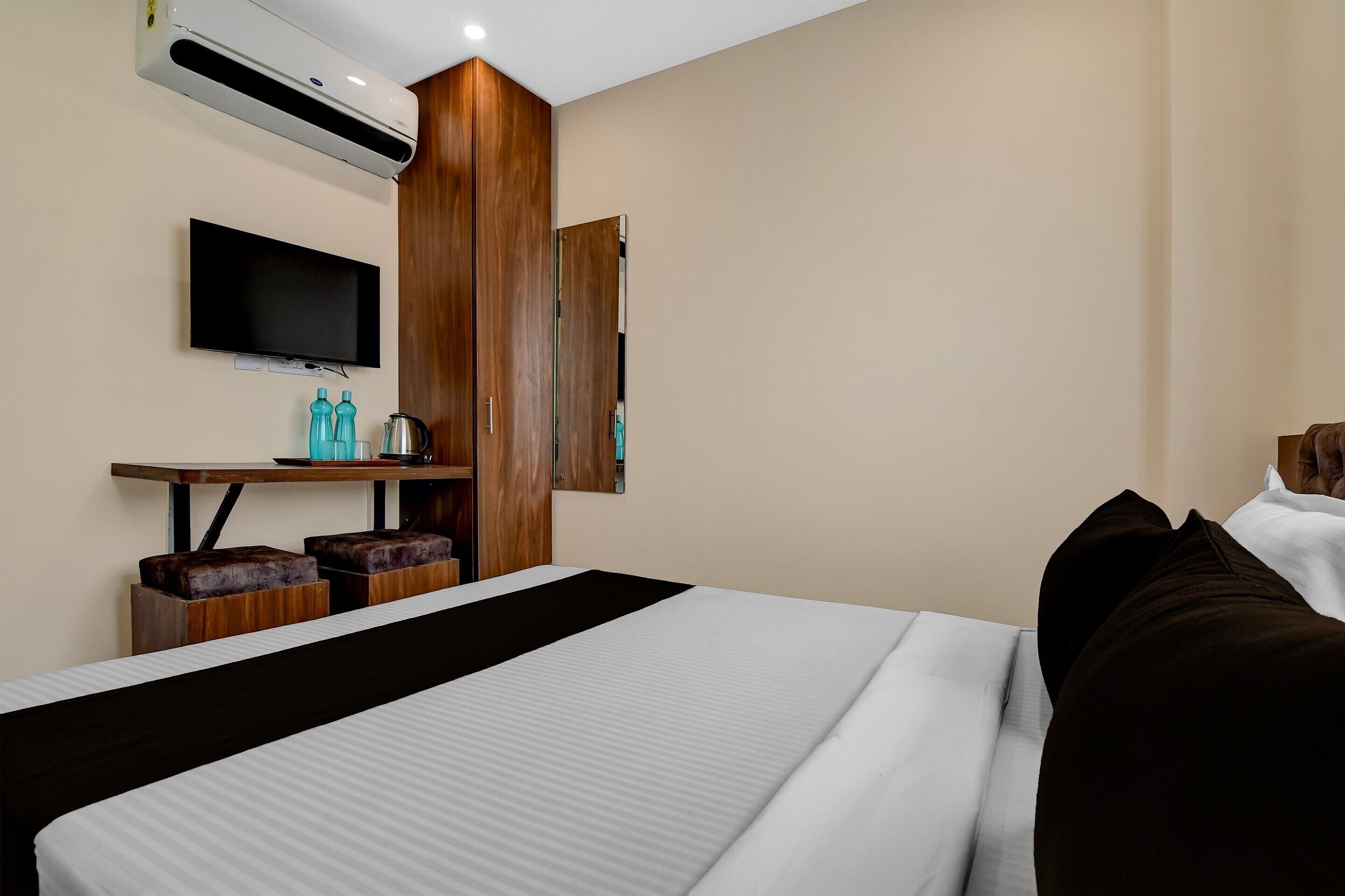 A hotel room with a bed, a TV, a wooden wardrobe, a desk with a kettle, and a mirror.