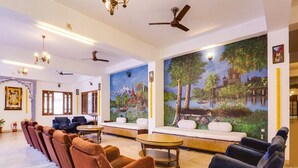 Un hall de hotel con una fila de sofás marrones y azules, un mural grande, ventiladores de techo y una decoración de estilo tradicional.