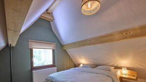 A bedroom with a sloped ceiling, a window with a view, a bedside table with a lamp, and a bench. - Beautiful cozy home in a wooded holiday park. (Rekem)