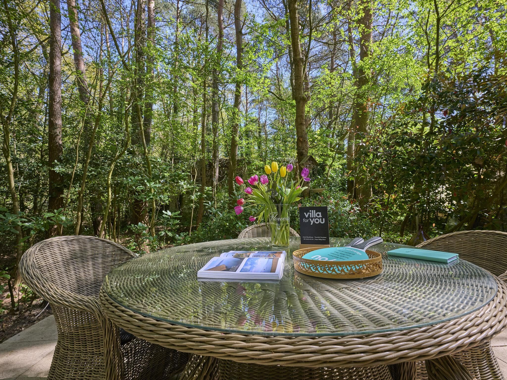 Una mesa de vidrio redonda con sillas de mimbre, un jarrón con flores y un letrero que dice 'villa for you' en un entorno exterior exuberante y verde.