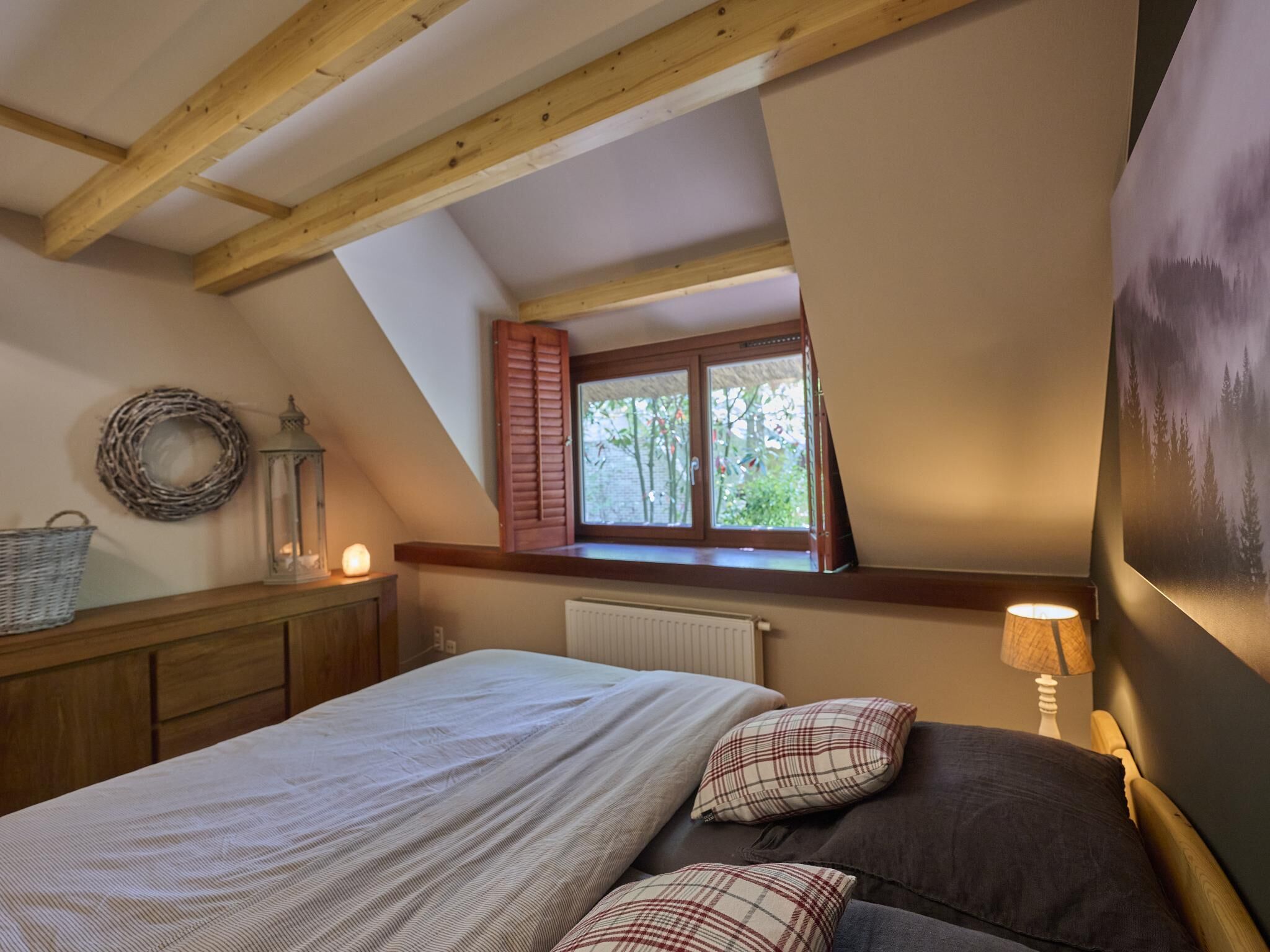 Una camera da letto con soffitto a falde, un letto con cuscini, un comodino, una lampada, una finestra con persiane e un termosifone a parete.