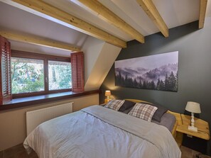 A bedroom with a large bed, a window with shutters, a painting of a forest, and a nightstand with a lamp.