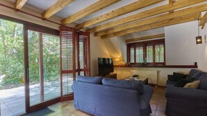 A living room with a sofa, a television, and a window with shutters. - Beautiful cozy home in a wooded holiday park. (Rekem)