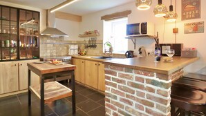 A modern kitchen with a central island, a stove, a microwave, and hanging utensils. - Charming cottage with a lovely terrace & garden (Gimnee)