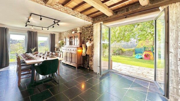 A dining area with a table set for four, a kitchen with stone walls, and a sliding glass door leading to a garden. - Charming cottage with a lovely terrace & garden (Gimnee)