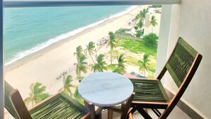 Een balkon met twee houten stoelen en een kleine tafel, met uitzicht op een strand met palmbomen en een stadsachtergrond in de verte.