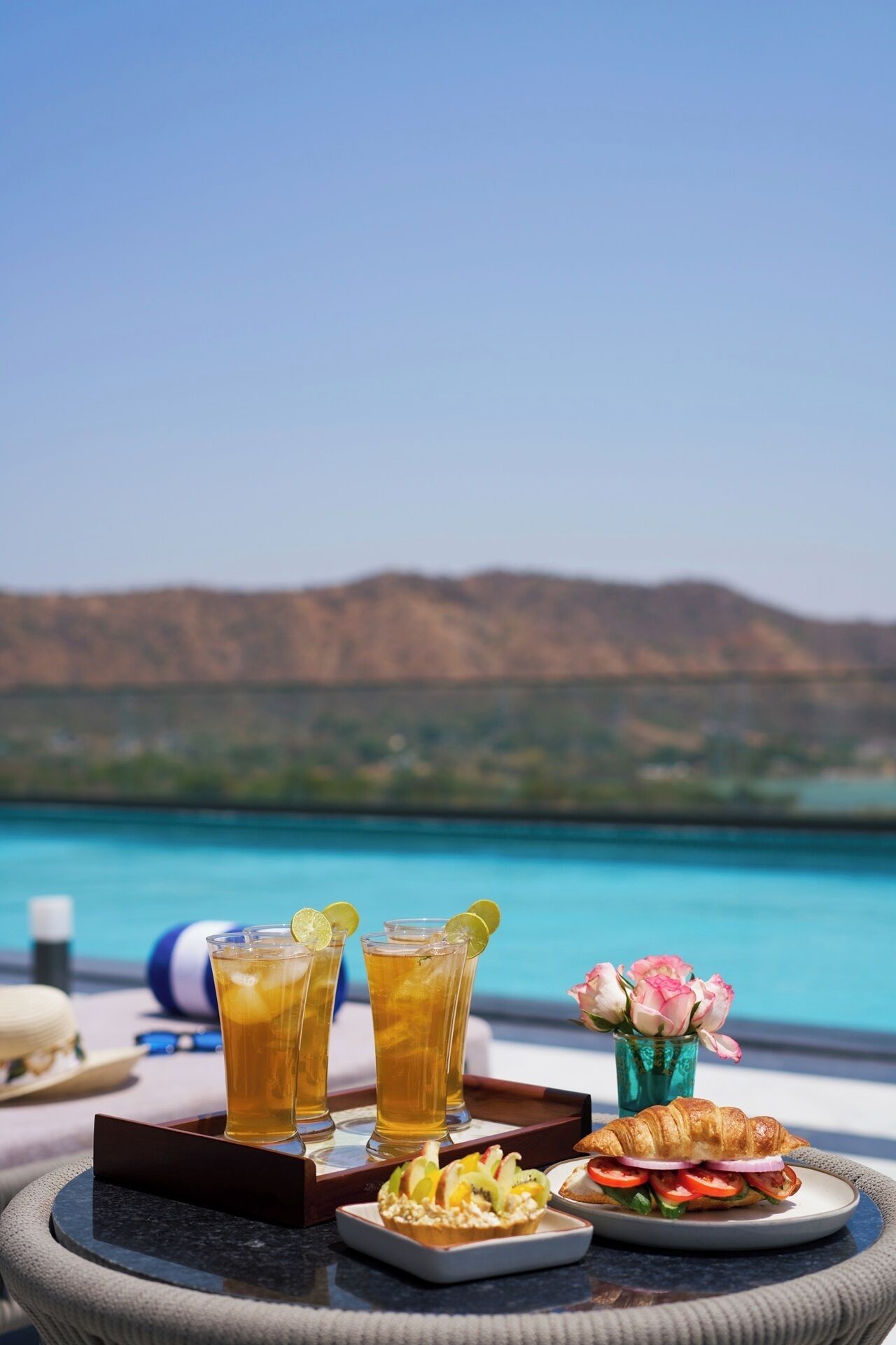 Una mesa con bebidas, un croissant y un plato de fruta en una terraza con vistas a un lago y montañas.