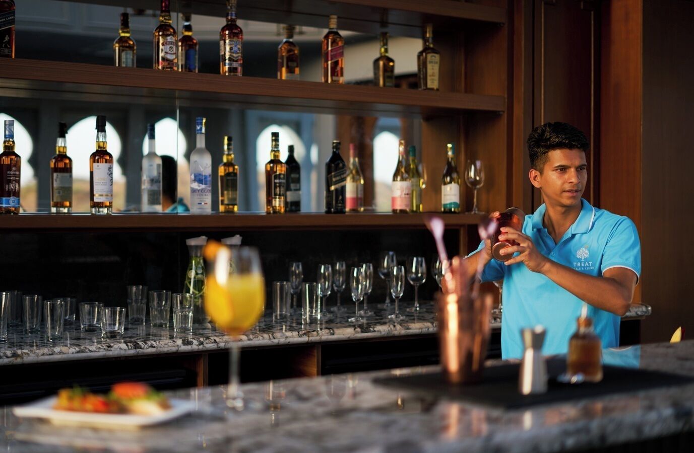 A bartender preparing a drink at a bar with a variety of bottles and glasses in the background.