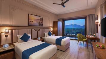 Habitación de hotel con dos camas, un escritorio, una silla, televisión y balcón con vistas a la montaña.