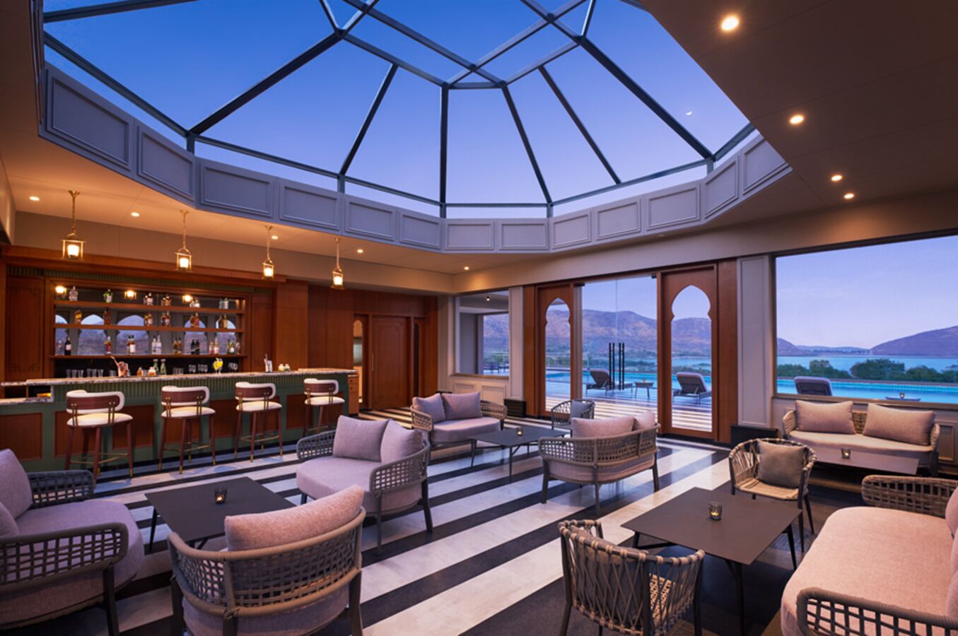A spacious indoor area with a bar, seating arrangements, and a large window offering a view of a lake and mountains.