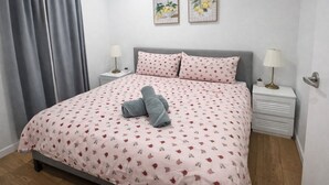 Ein Schlafzimmer mit einem Bett, zwei Nachttischen, einer Lampe auf jedem Tisch sowie einem grauen Vorhang und zwei gerahmten Bildern an der Wand.