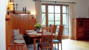 Ein Essbereich mit einem Holztisch, Stühlen und einer Bank. Ein Fenster mit Vorhängen, eine Holzbalkendecke und ein Teppichboden sind ebenfalls vorhanden.