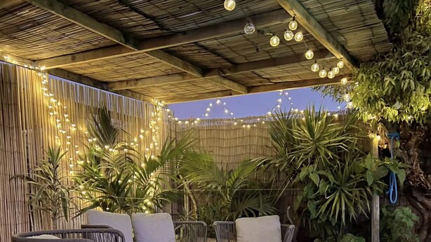 Un patio al aire libre con muebles de mimbre, luces colgantes y plantas en macetas.
