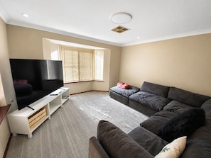 A living room with a flat-screen TV, a grey sofa, a white entertainment unit, and a large window with blinds.
