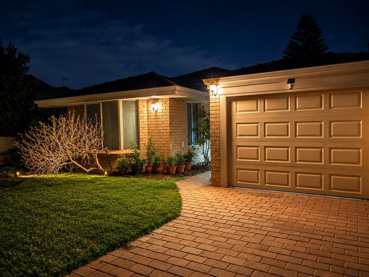 A residential garage with a brick pathway leading to it, surrounded by a well-maintained lawn and potted plants.