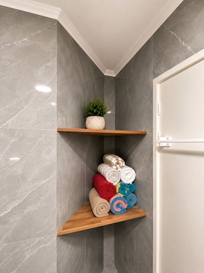 A corner of a room with grey marble walls, a wooden shelf with rolled towels, and a small potted plant. - Perth Beach House (Mullaloo)