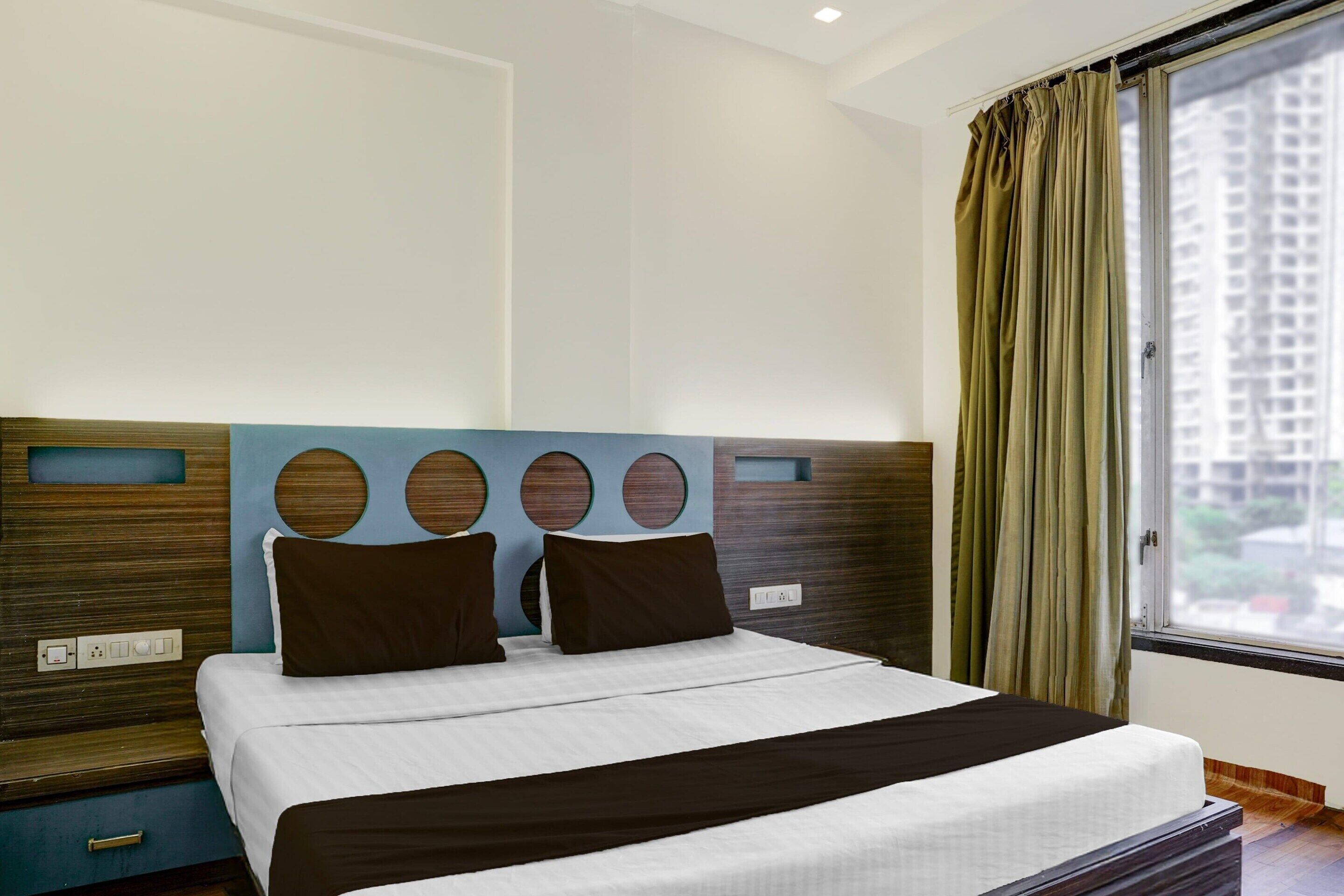A hotel room with a bed, a window with curtains, a wall with circular cutouts, and a power outlet.