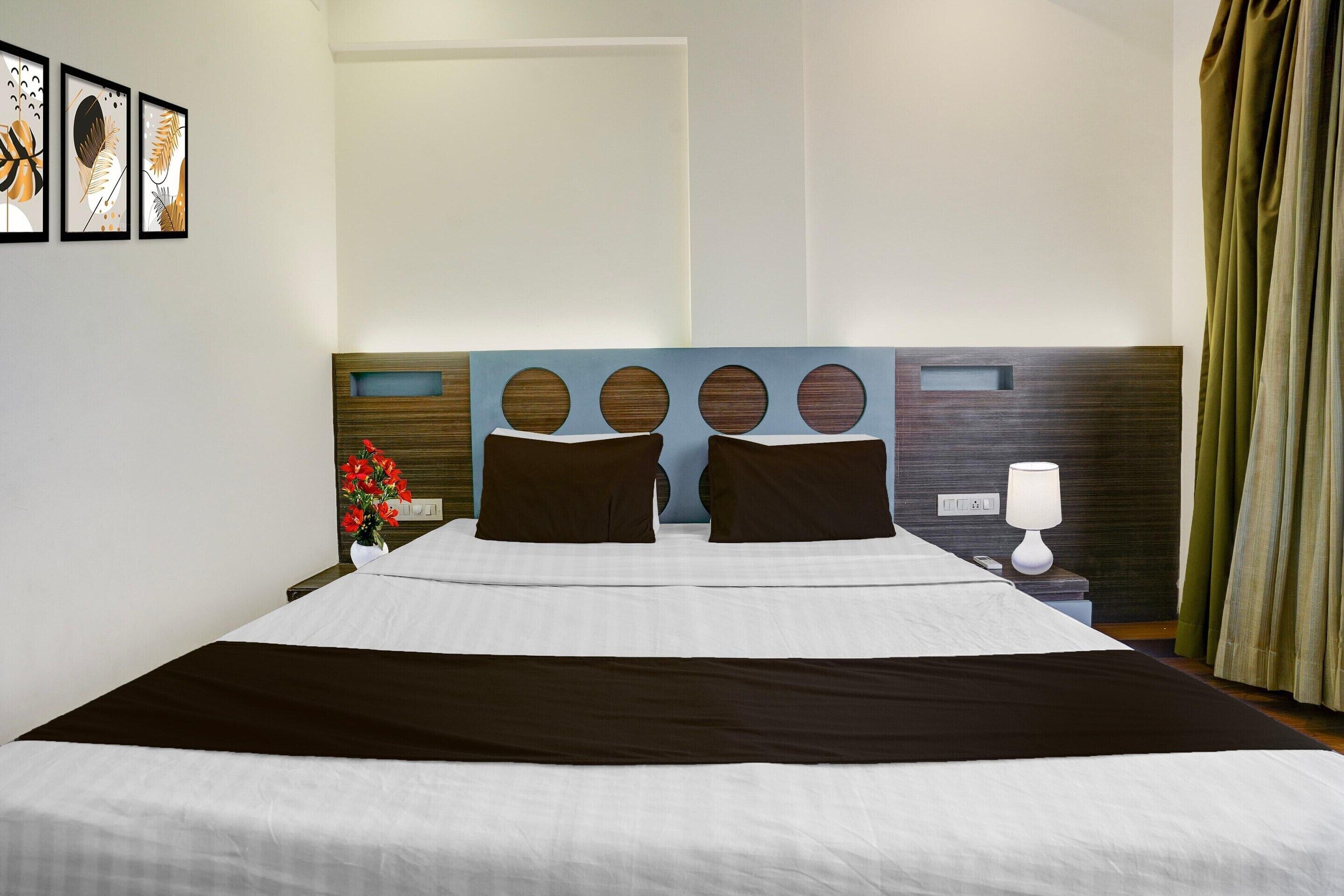 A hotel room with a large bed, a headboard with circular cutouts, a bedside table with a lamp, and a wall with framed artwork.