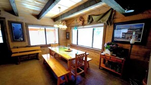 Een houten interieur met een lange tafel, banken, grote ramen en een klein kitchenette.