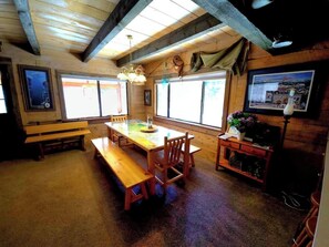 A wooden interior with a long table and benches, large windows, and a small kitchenette. - The Perfect Cozy Cabin of Elo Road! (McCall)