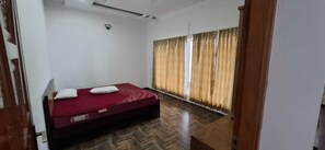 A room with a bed, a security camera, a window with curtains, and a herringbone-patterned floor.