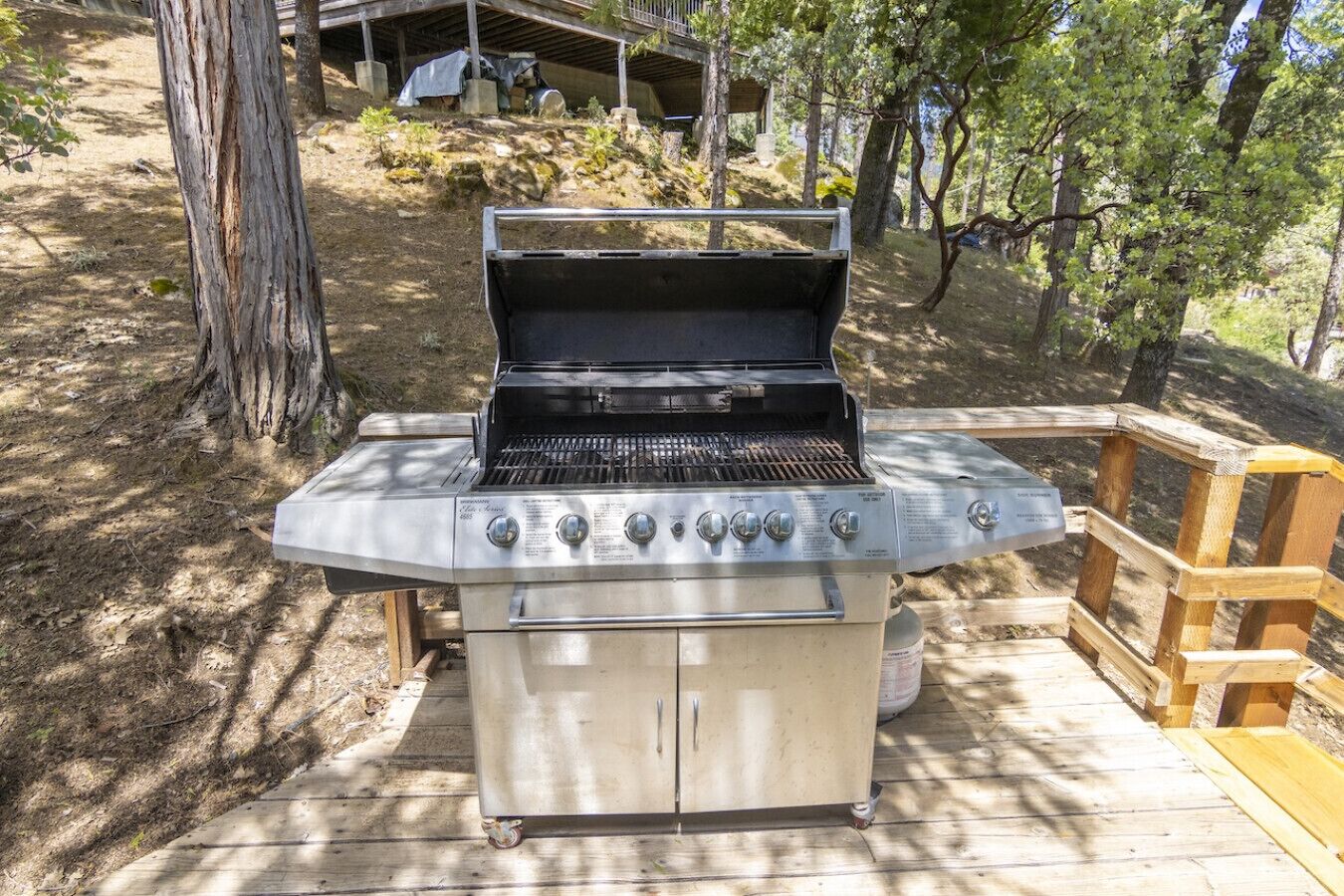 A portable gas grill with four burners, a side shelf, and a storage cabinet, set on a wooden deck in a wooded area.