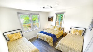 A room with three beds, each with a different style of bedding. There are windows with wooden railings, and the room has a wooden floor.