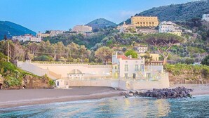 A coastal building with a beach in the foreground, a rocky outcrop, and a hillside with buildings in the background. - Villa Sirenea: A splendid and luminous three-story villa located right on the beach, with Free WI-FI. (Vico Equense)