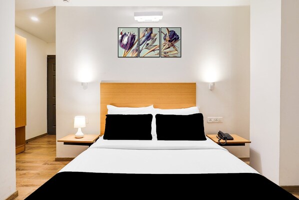 A hotel room with a wooden headboard, a bed with white and black bedding, a bedside table with a lamp, a phone, and a wall-mounted artwork. - Super BTM Layout Madiwala Formerly Rooms (Bengaluru)