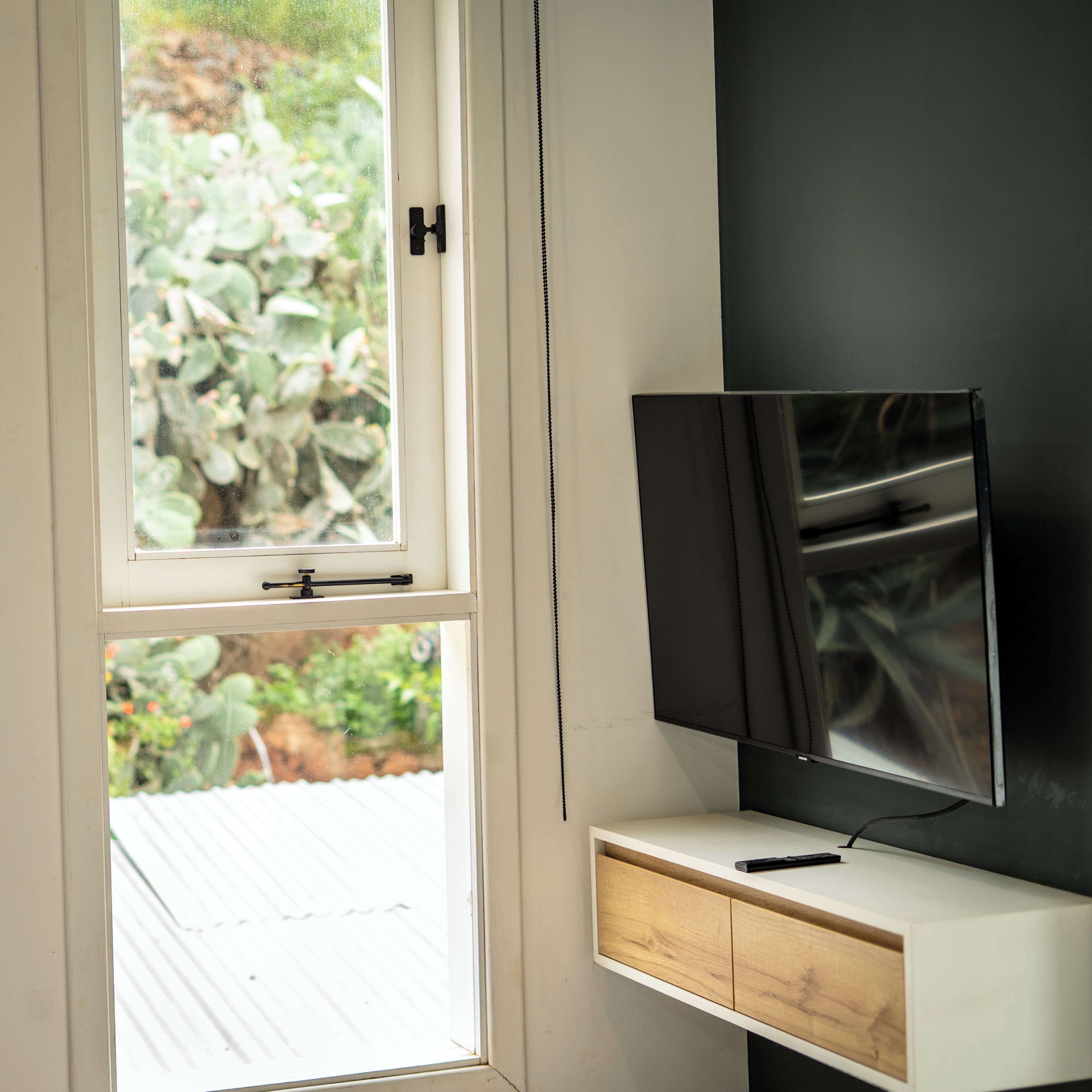 A room with a television mounted on a wall, a window with a view of greenery, and a white cabinet with drawers.