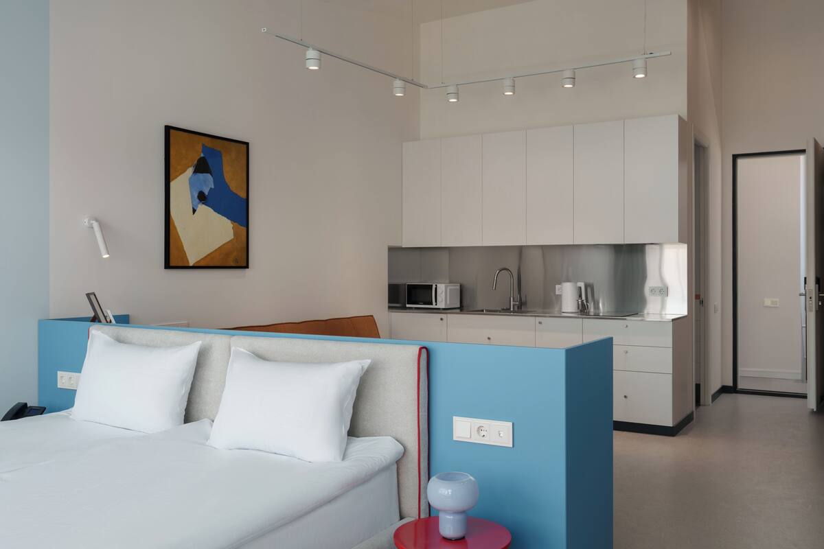 Una habitación de hotel moderna con cama, una kitchenette pequeña, un cuadro en la pared y una pared de acento azul.